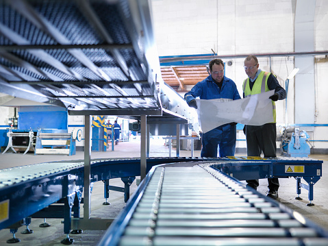 Two workers reviewing plans beside a roller conveyor line on a factory floor